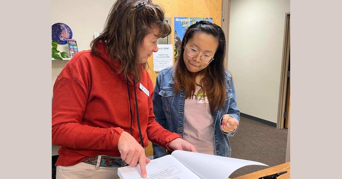 Teacher points at a page of an open book as a student stands next to her.